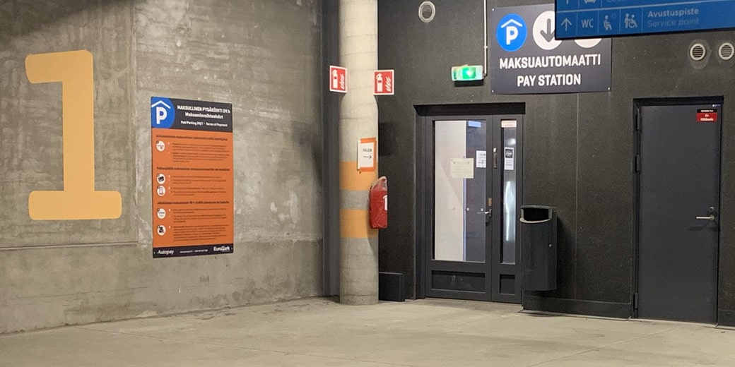 P-Matkakeskus Kuopio, kulmaus parkkitalon sisällä, tasolla P1, jossa on ohjekyltti ja ovi, jonka yläpuolella lukee