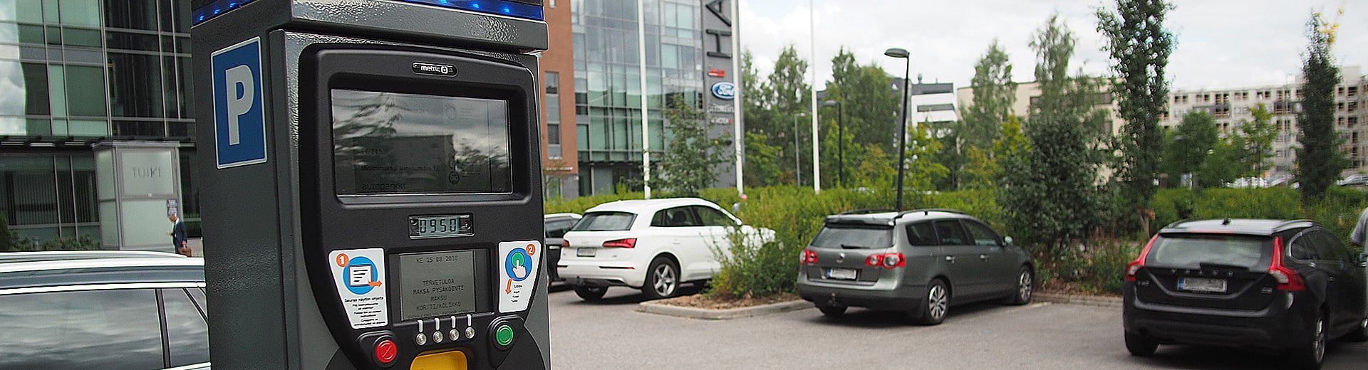 Metric maksuautomaatti, taustalla parkkipaikka, autoja ja rakennuksen seinää.