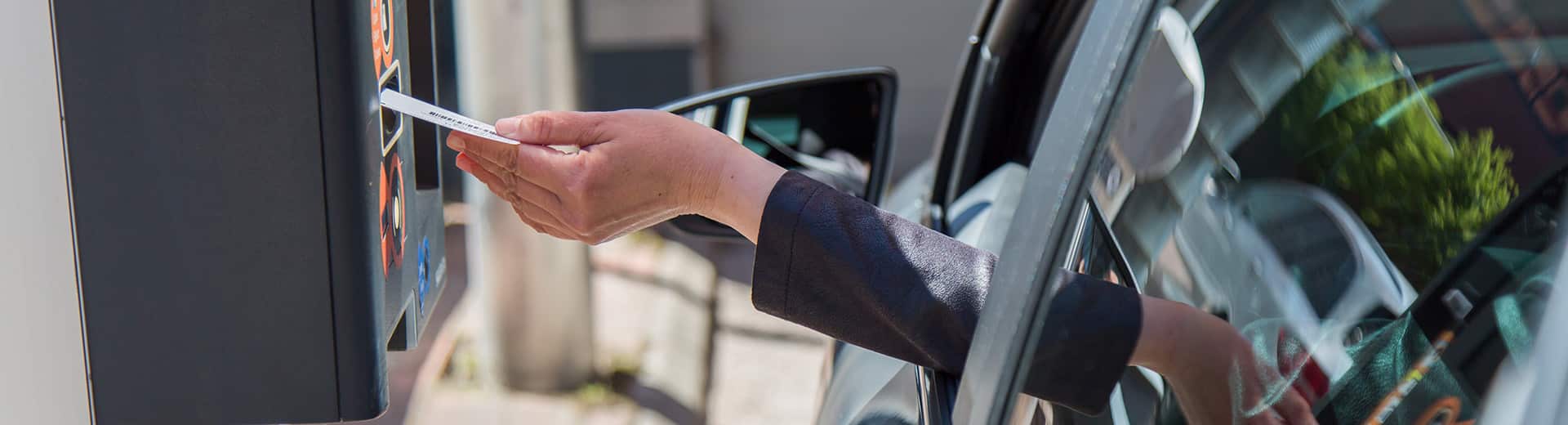 Valokuva, jossa näkyy henkilön käsi, joka ojentuu autosta laittamaan parkkilippua automaattiin.